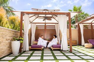Two people in white robes relax under a cabana, enjoying drinks in a serene outdoor setting surrounded by greenery and decorative elements.